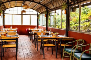 Un restaurant avec des tables et des chaises en bois, un mur rouge et de grandes fenêtres donnant sur un espace verdoyant.