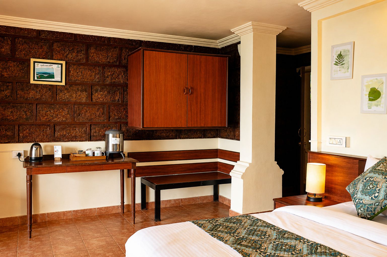 Une chambre d’hôtel avec un banc en bois, un lit recouvert d’une courtepointe à motifs, une machine à café et un certificat encadré accroché au mur.