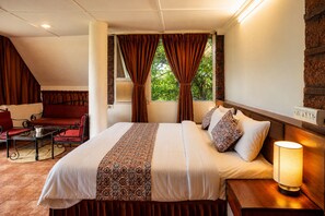 A hotel room with a large bed, a bedside table with a lamp, a window with curtains, a seating area with a sofa and chairs, and a view of greenery outside. - Wind Chalet (Patan)