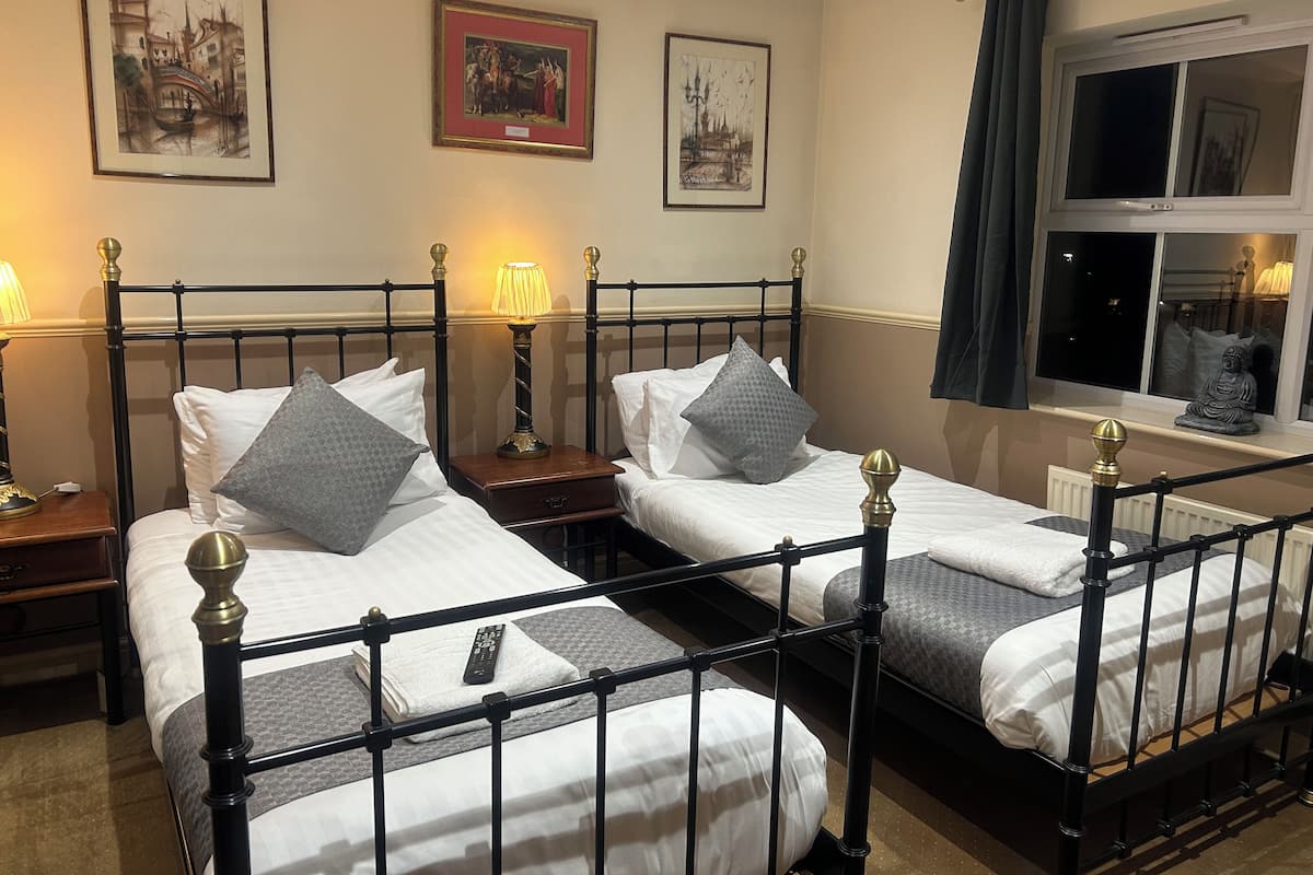 A hotel room with two beds, each with a nightstand, a lamp, and a remote control on the bed. There are framed pictures on the wall and a window with a view of the outside.