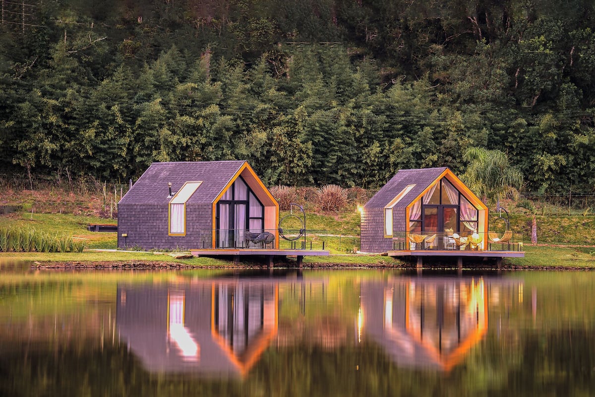 Deux maisons flottantes modernes, construites en bois, dotées de grandes fenêtres et de balcons, et situées sur un lac paisible, avec une forêt dense en arrière-plan.