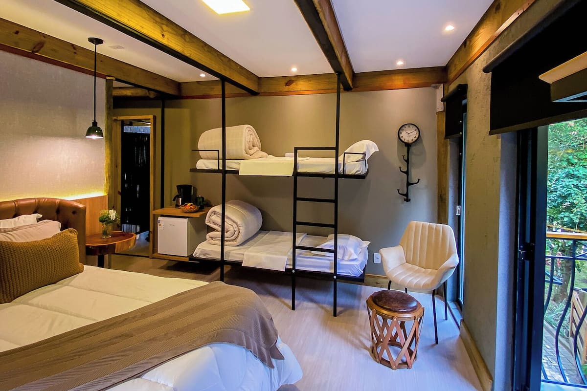Une chambre d’hôtel avec un grand lit, un lit plus petit, un bureau avec une chaise, une petite table, une lampe, une horloge et une fenêtre donnant sur un paysage verdoyant.