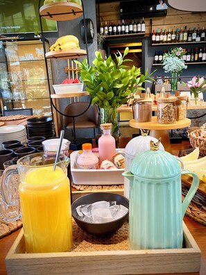 Un buffet de petit-déjeuner avec divers aliments, une carafe de jus d’orange, un bol de glaçons et une théière sur un plateau en bois.