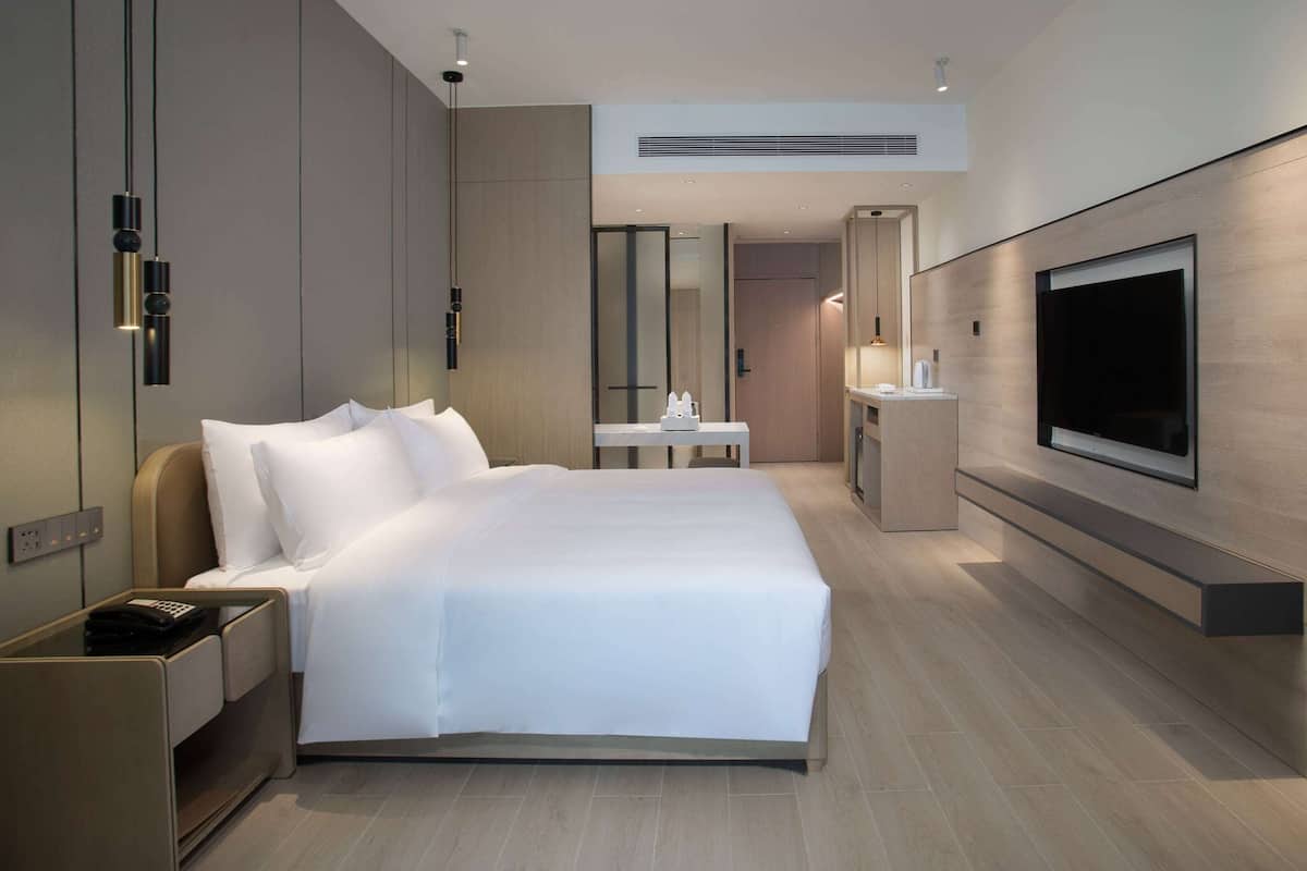 A modern hotel room with a large bed, bedside table, TV, and a small desk with a chair.