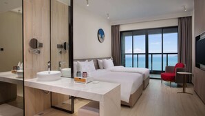 A modern hotel room with a large bed, a separate sofa, a round table, a large mirror, and a view of the sea.