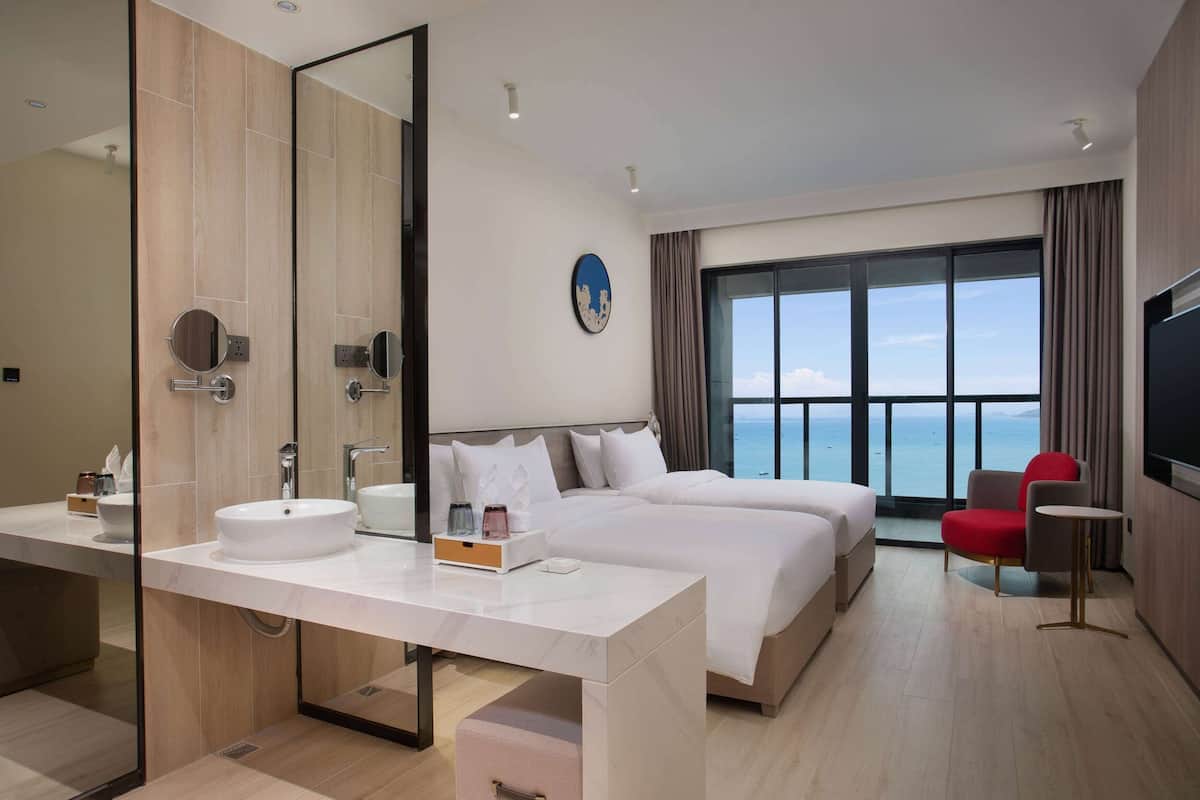 A modern hotel room with a large bed, a separate sofa, a round table, a large mirror, and a view of the sea.