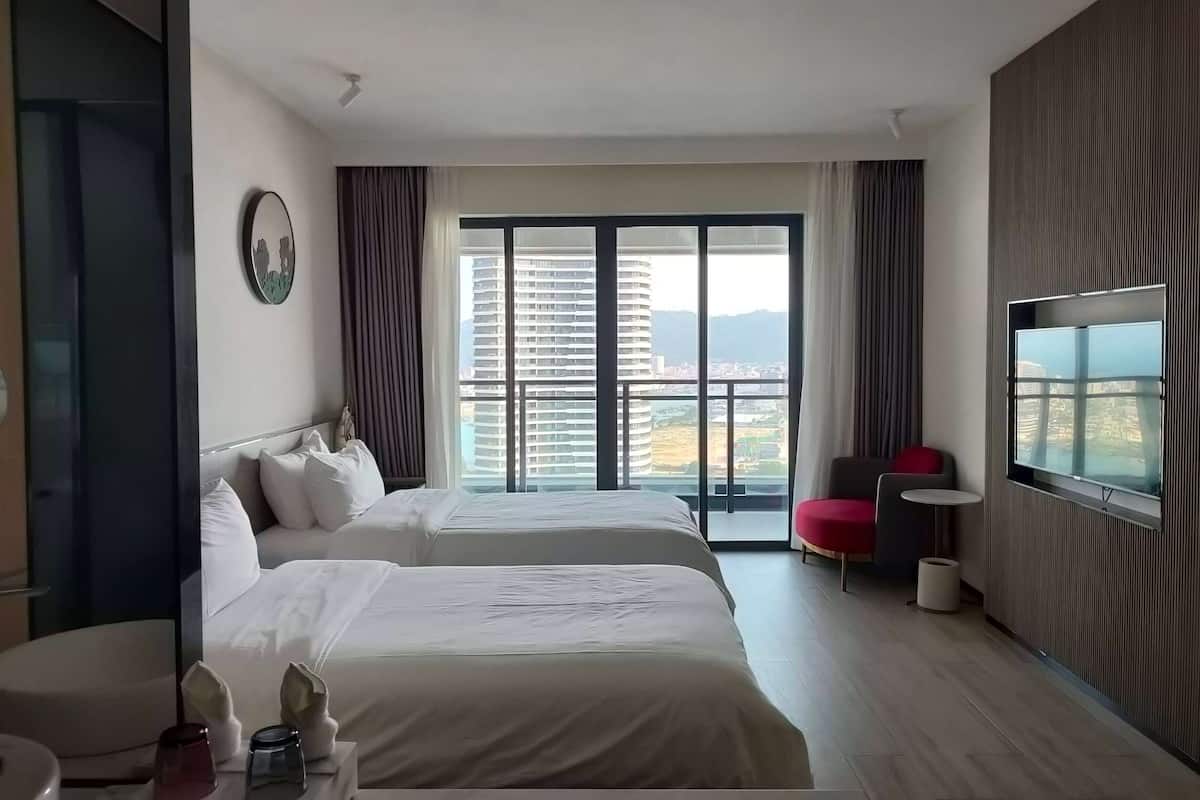 A hotel room with two beds, a TV, a balcony with a view of buildings and mountains, and a red chair.