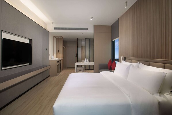A modern hotel room with a large bed, a flat-screen TV mounted on the wall, a desk with a chair, and a small bathroom area.