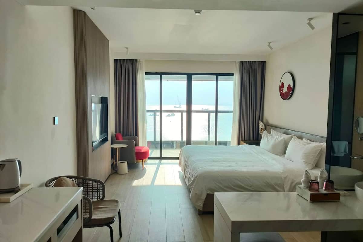 A hotel room with a bed, a desk, a chair, a small table, and a view of the sea through large windows.