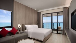 A modern hotel room with a large bed, a sofa, a round coffee table, and a balcony with a view of the sea.