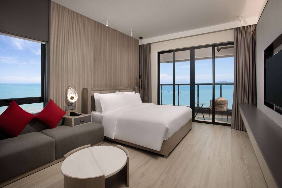 A modern hotel room with a large bed, a sofa, a round coffee table, and a balcony with a view of the sea.