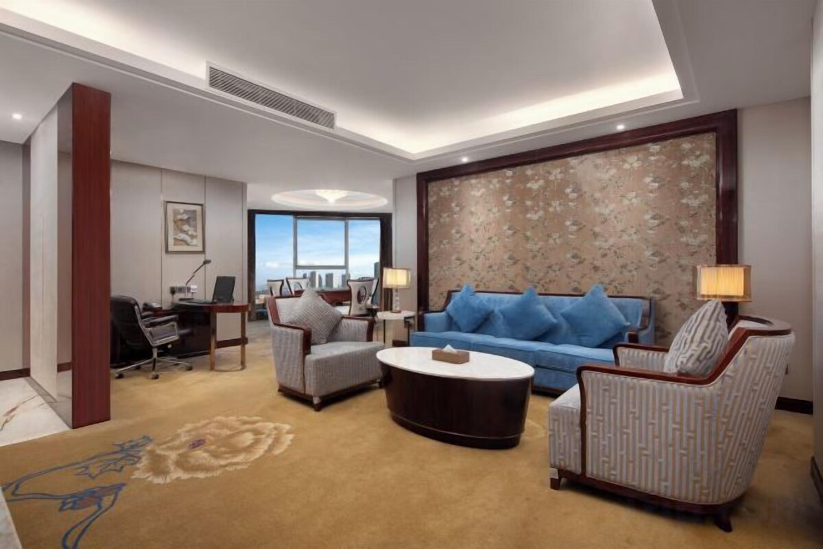 A hotel room with a sofa, armchairs, a coffee table, a desk, and a large window.