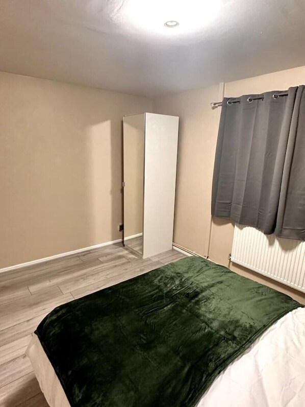 A bedroom with a bed, a green blanket, a white radiator, and a grey curtain.