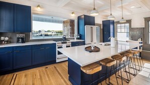 A modern kitchen with dark blue cabinets, a large island with stools, stainless steel appliances, and pendant lighting. - The Capone House | Historic Luxury on the Bay (Atlantic City)