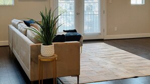 A living room with a beige sofa, a small side table with a plant, and a large window with blinds. - 3-Story Downtown Houston Safe Haven (Houston)