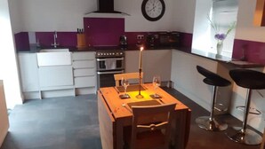 A compact kitchen with a dining table, two bar stools, a clock, and a window with blinds.