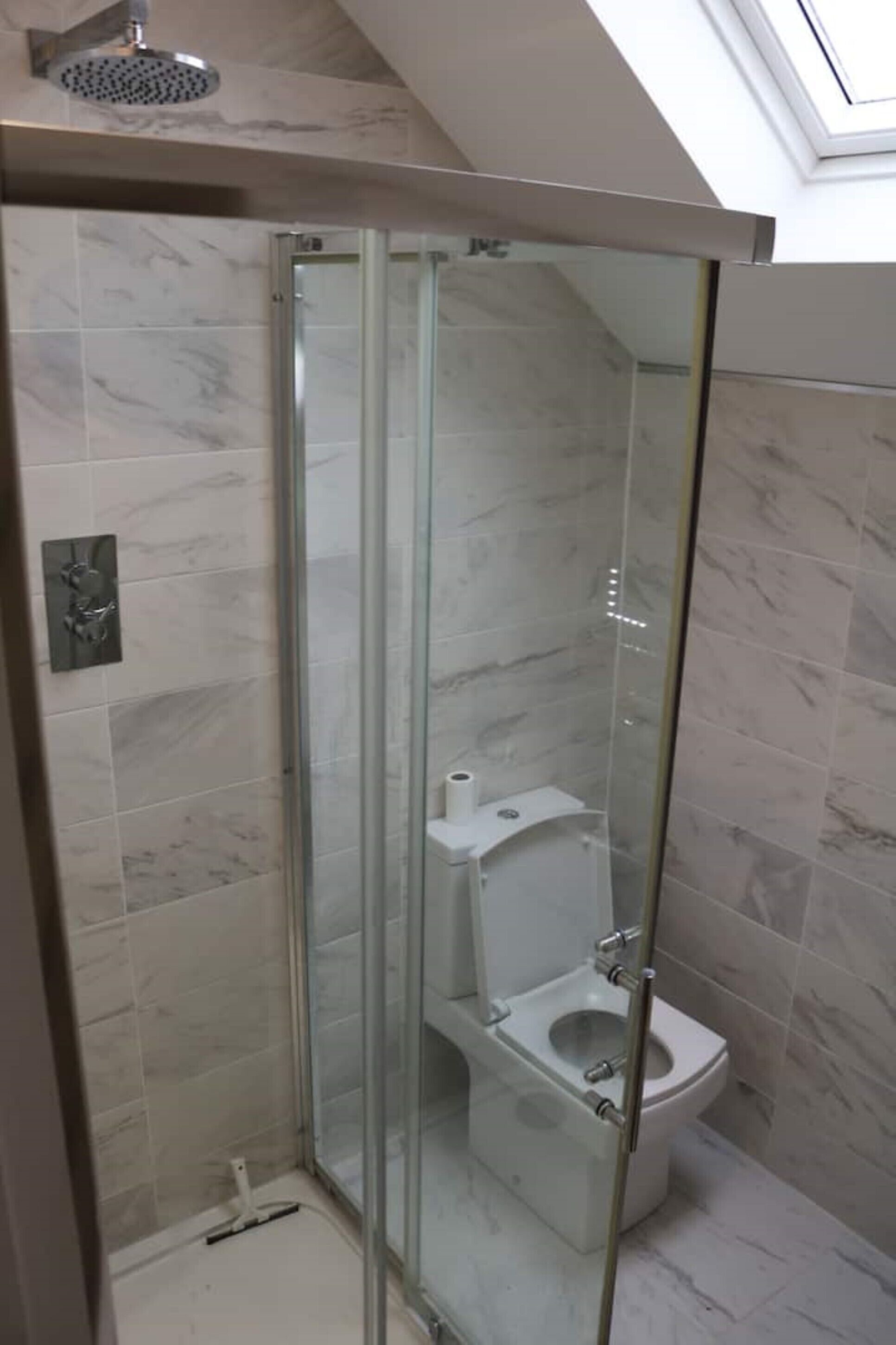 A glass-enclosed shower stall with a toilet inside, marble-tiled walls, and a skylight above.