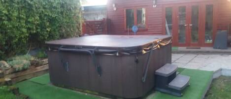 A hot tub in a backyard with a wooden deck and a red building in the background.