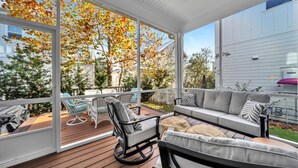 Un patio cubierto con terraza de madera, mobiliario exterior y ventanas amplias que ofrecen vistas a árboles y una casa.