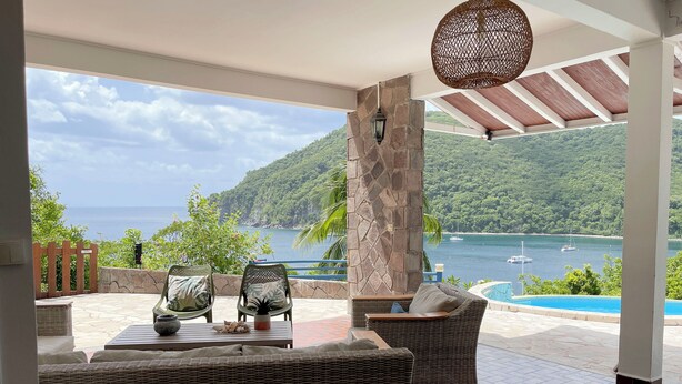 A terrace with wicker furniture, a stone pillar, and a view of the sea and mountains.