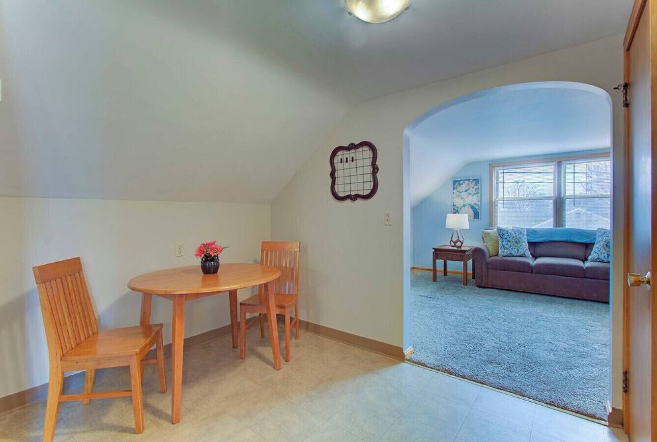 A room with a dining table and chairs, a small wall decoration, and a view into a living area with a couch and a window.