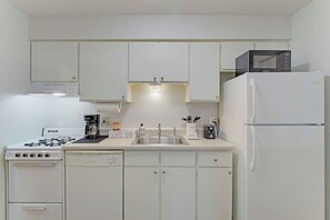 Une cuisine de taille réduite avec des meubles blancs, une cuisinière à gaz, une machine à café, un four à micro-ondes et un réfrigérateur.