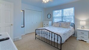 Quarto com cama, ventoinha no teto, janela, mesa de cabeceira e porta.