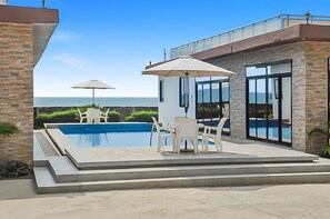 A modern outdoor pool area with a view of the ocean, a covered seating area, and a building with large windows. - 2 Bed/2.5 Bath Villa (Lewa) (Monrovia)