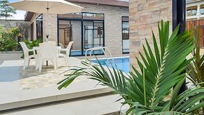 An outdoor pool area with white plastic chairs and a large umbrella, surrounded by stone walls and potted plants.