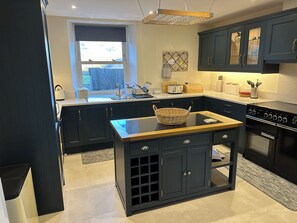 A modern kitchen with dark cabinetry, a central island, a built-in oven, and a window with a view of the outside. - Ings House (Hawes)