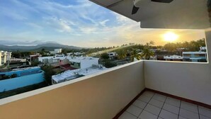 Uma varanda com ventilador de teto, piso de azulejos e vista para uma área residencial com casas e palmeiras ao pôr do sol.