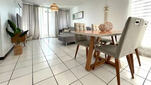 Uma sala de estar moderna com área de jantar, apresentando uma janela grande, um sofá cinza e uma mesa de jantar de madeira com cadeiras.