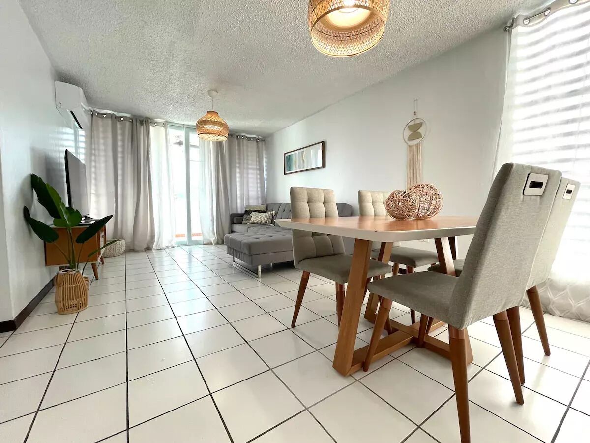 A modern living room with a dining area, featuring a large window, a grey sofa, and a wooden dining table with chairs.