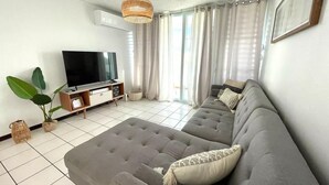 Uma sala de estar moderna com um sofá cinza, uma televisão de tela plana e uma janela grande com cortinas.