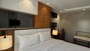 A modern hotel room with a large bed, a wooden headboard, a TV mounted on the wall, a bedside table with a lamp, and a small seating area with a bench. - Luxury Abode Near Walking Street

 (Malabañas)