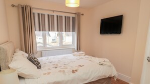 A bedroom with a bed, a TV mounted on the wall, a window with curtains, a bedside table with a lamp, and a wooden floor. - 55 Wellwood Street (Morpeth)
