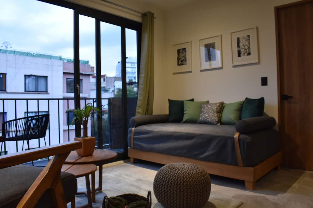 A modern bedroom with a sofa bed, a small table, and a chair. There are three framed pictures on the wall and a large window with a view of buildings outside.