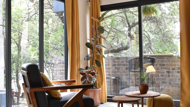 A modern living room with a large window, a wooden chair, a small table, and a potted plant. - Apartments Art Tanat (Mexico City)