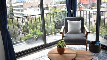 Un balcón con vistas a la ciudad, un sillón gris con un cojín blanco y una pequeña mesa de madera con una planta en maceta.