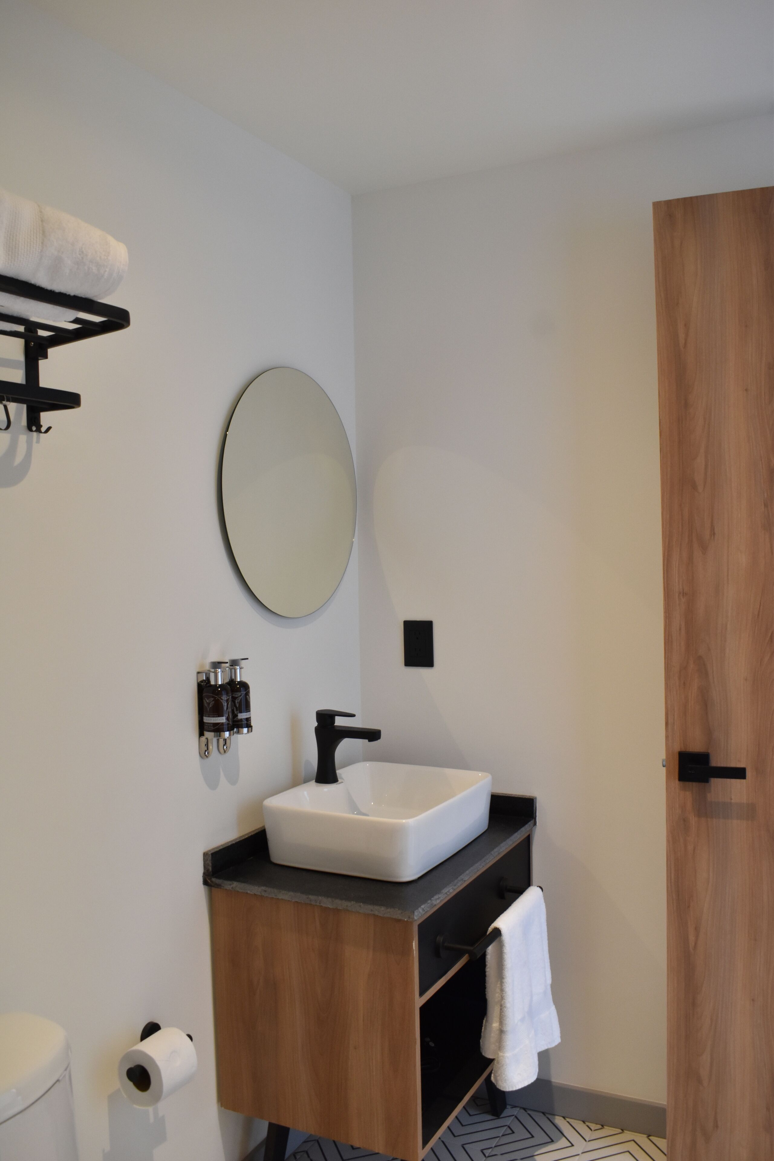 Ein modernes Badezimmer mit einem weißen Waschbecken, einem schwarzen Wasserhahn und einem runden Spiegel. Darüber befindet sich ein Handtuchhalter und an der Wand ein Toilettenpapierhalter.