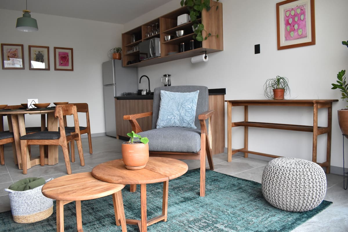 A modern living room with a dining area, wooden furniture, a grey sofa, a round wooden coffee table, and a green rug.