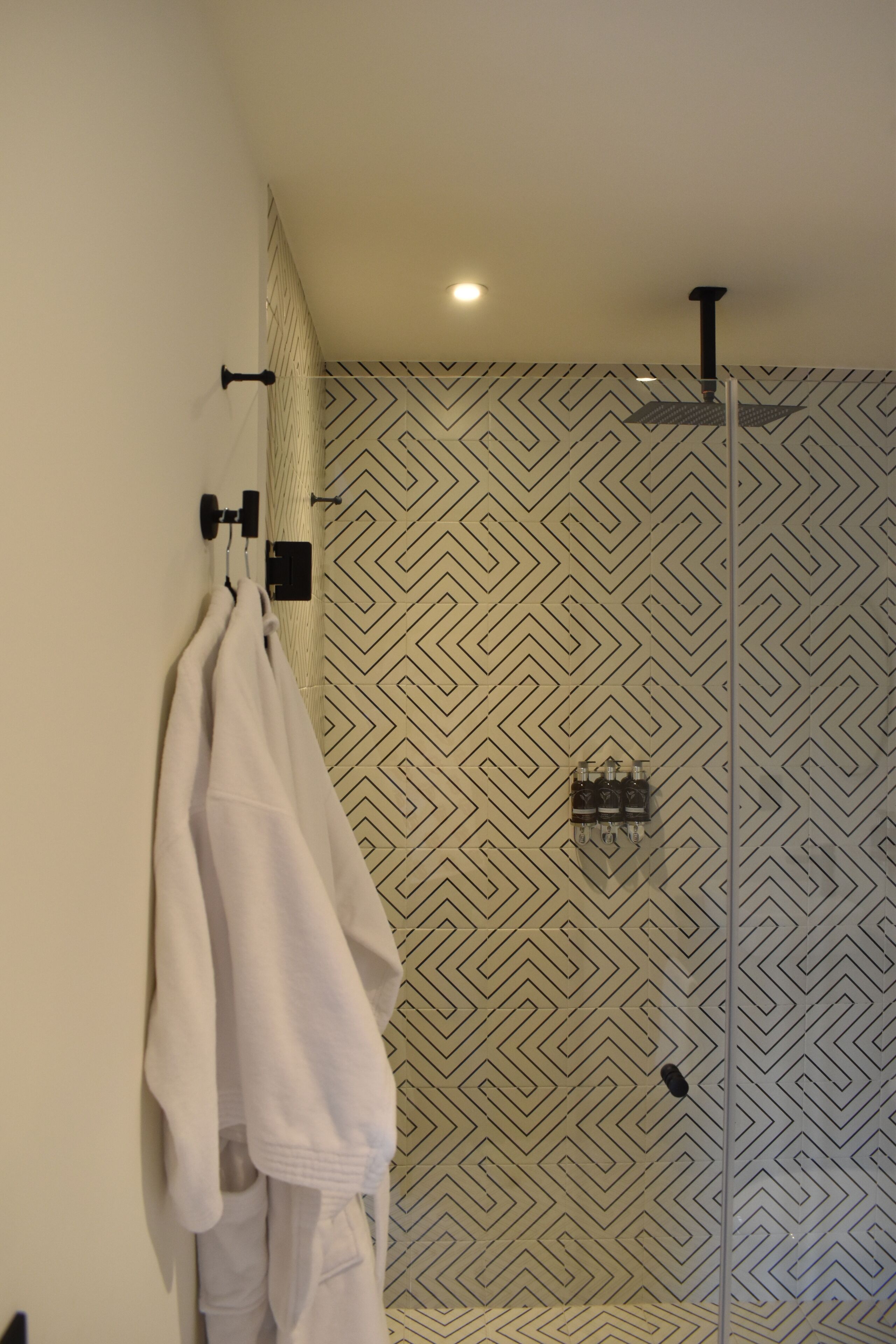Un baño con pared de diseño geométrico, un perchero con toallas y una zona de ducha con luz en el techo.