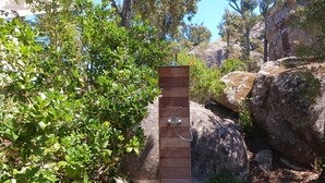 Una ducha exterior de madera con manguera metálica, frente a un fondo de rocas y follaje.