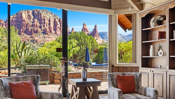Eine Terrasse mit zwei gemusterten Sesseln, einem kleinen Tisch und einer Steinskulptur. Ein Holzregal und ein großes Fenster mit Blick auf rote Felsformationen und Grünflächen sind ebenfalls zu sehen.