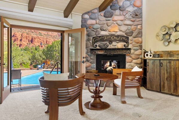 A stone fireplace with a sign above it, a pool visible through an open door, and a wooden table with chairs in the foreground.
