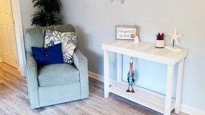 A living room with a grey armchair, a white console table, and wall decorations including a 'Fresh Seafood' sign, a turtle sign, and a framed picture with a message.