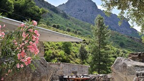 Un patio al aire libre con un muro de piedra, una mesa redonda de madera y sillas. Se divisa una montaña con cumbre al fondo.