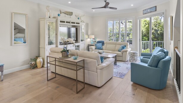 Un salon spacieux avec un canapé beige, un fauteuil bleu et une table basse. On aperçoit un téléviseur posé sur un meuble et un ventilateur de plafond.