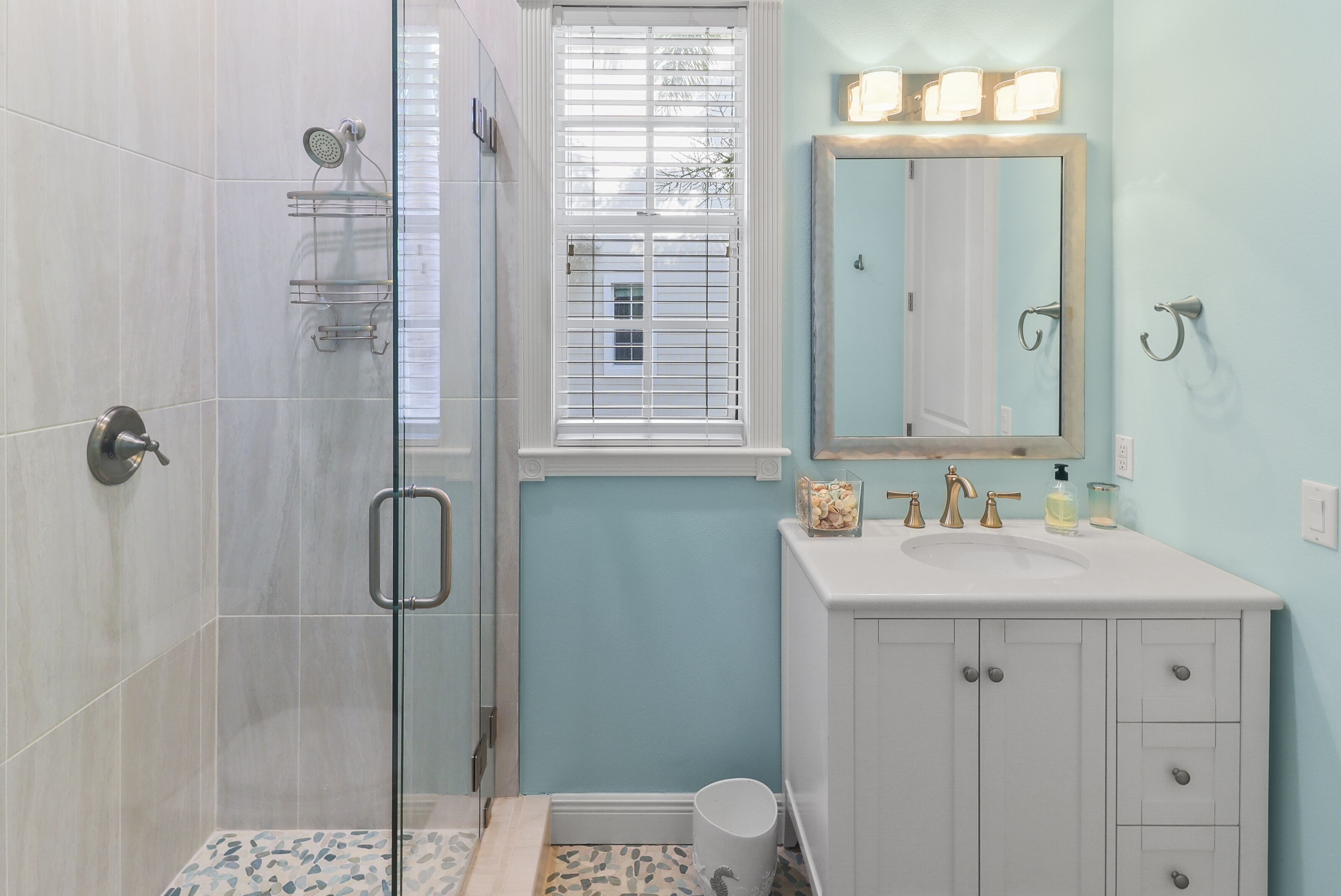 A bathroom with a glass shower enclosure, a white sink with gold fixtures, and a mirror with lights above.