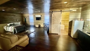 A spacious living room with a sofa, a coffee table, and a bed with a quilt. There is a kitchen area with a refrigerator and a sink. - Newly added studio apartment, above garage.  (Marvell)